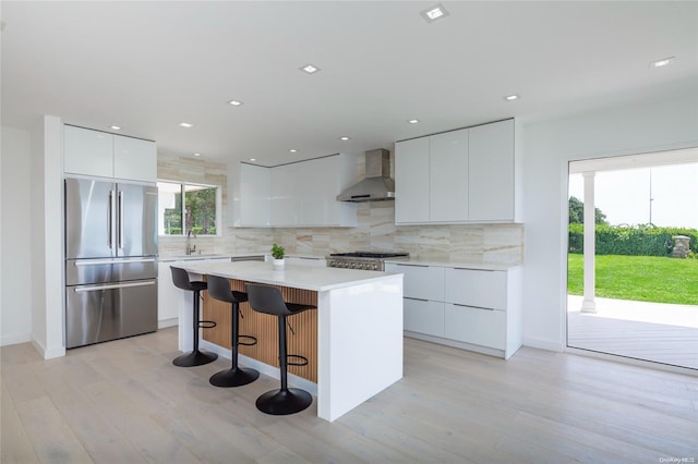 kitchen with wall chimney exhaust hood, light wood-type flooring, a kitchen island, white cabinetry, and stainless steel appliances