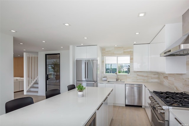 kitchen with white cabinets, wall chimney range hood, sink, light wood-type flooring, and premium appliances