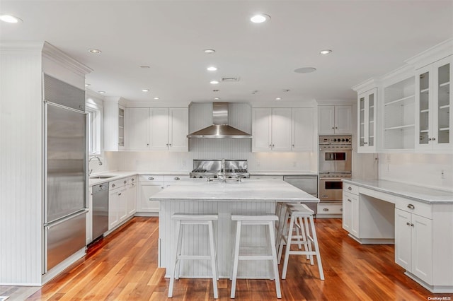 kitchen featuring wall chimney range hood, light hardwood / wood-style flooring, a kitchen bar, a kitchen island, and appliances with stainless steel finishes