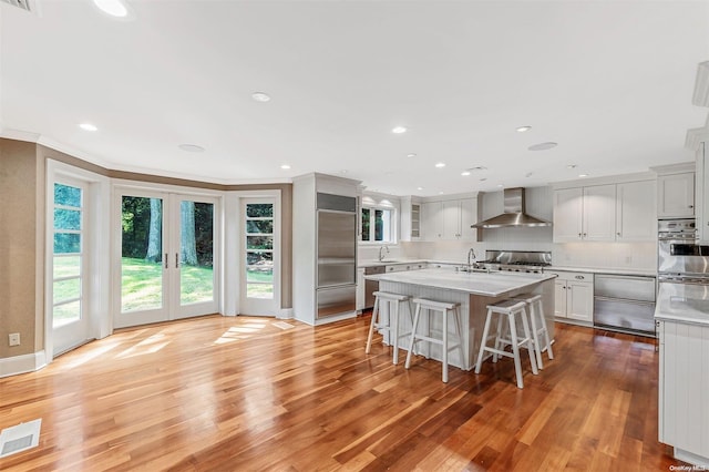 kitchen with a breakfast bar, a kitchen island with sink, crown molding, light hardwood / wood-style flooring, and wall chimney exhaust hood