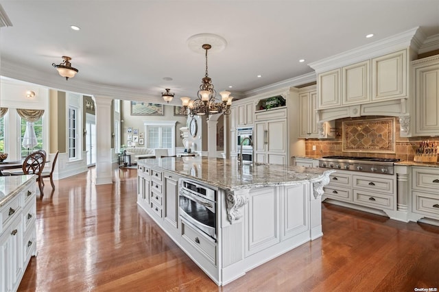 kitchen featuring appliances with stainless steel finishes, decorative columns, a kitchen island with sink, light hardwood / wood-style flooring, and hanging light fixtures