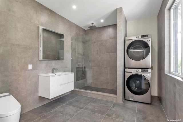 clothes washing area with dark tile patterned flooring, sink, tile walls, and stacked washer and clothes dryer