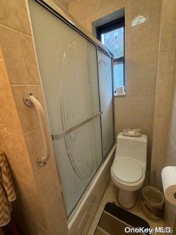 bathroom with combined bath / shower with glass door, toilet, and tile walls