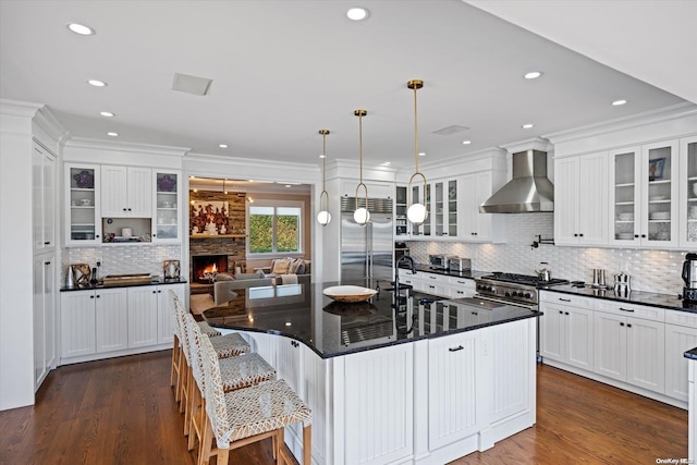 kitchen with dark wood-type flooring, a center island with sink, stainless steel built in fridge, hanging light fixtures, and wall chimney exhaust hood