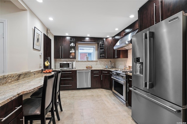 kitchen with wall chimney exhaust hood, dark brown cabinetry, a breakfast bar area, and appliances with stainless steel finishes