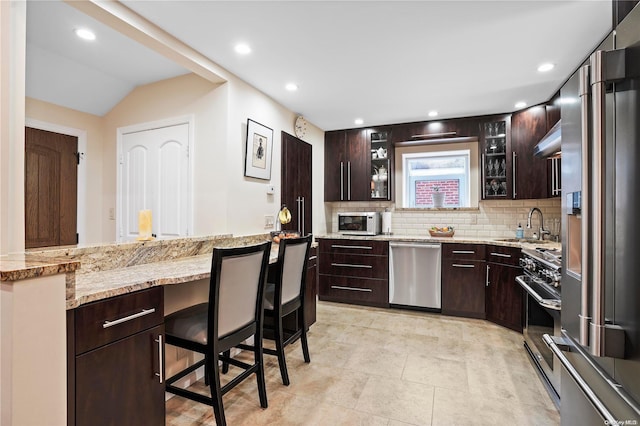 kitchen featuring a kitchen bar, decorative backsplash, light stone countertops, dark brown cabinetry, and stainless steel appliances