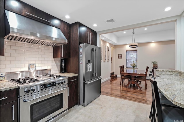 kitchen featuring decorative backsplash, premium appliances, wall chimney range hood, pendant lighting, and light hardwood / wood-style flooring