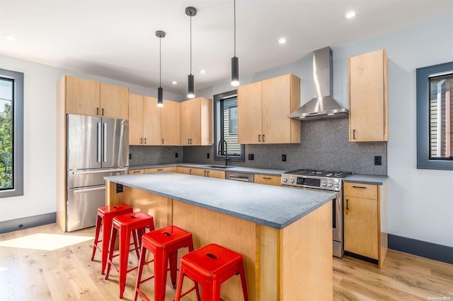 216 N St, Greenport, NY 11944 - kitchen featuring wall chimney range hood, appliances with stainless steel finishes, light brown cabinetry, a breakfast bar area, and light wood-type flooring