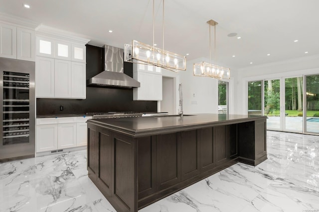 kitchen with beverage cooler, wall chimney range hood, decorative light fixtures, a center island with sink, and white cabinets