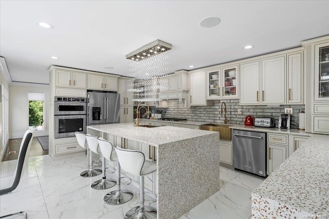 kitchen with tasteful backsplash, light stone counters, stainless steel appliances, sink, and a center island with sink