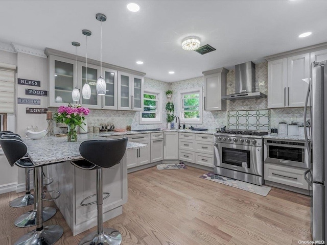 kitchen featuring a kitchen bar, stainless steel appliances, wall chimney range hood, white cabinets, and hanging light fixtures