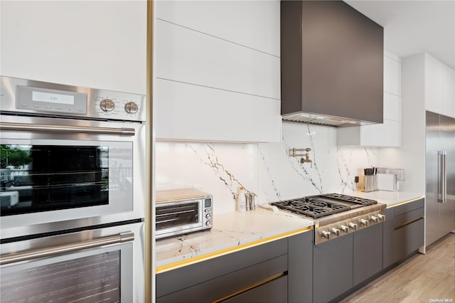 kitchen featuring light stone counters, stainless steel appliances, wall chimney range hood, white cabinets, and light hardwood / wood-style floors