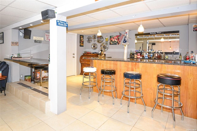 bar with a drop ceiling, sink, dark stone countertops, decorative light fixtures, and light tile patterned flooring