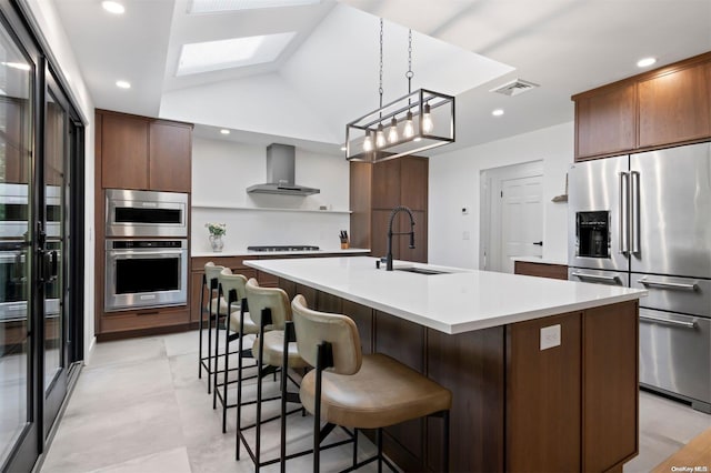 kitchen with appliances with stainless steel finishes, vaulted ceiling, a kitchen island with sink, sink, and wall chimney range hood