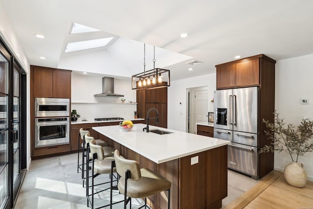 kitchen featuring a center island with sink, stainless steel appliances, light countertops, a sink, and wall chimney range hood