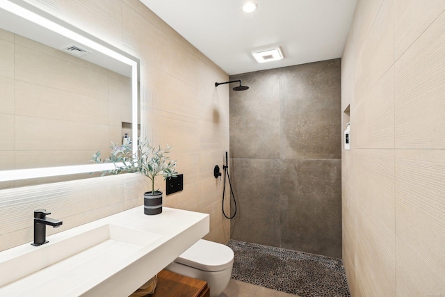 bathroom featuring visible vents, toilet, a sink, a tile shower, and tile walls