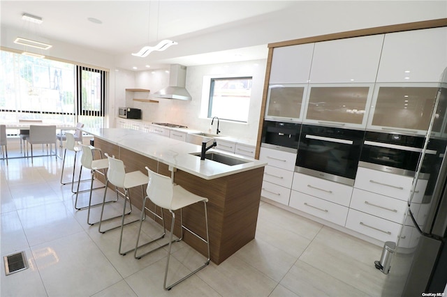 kitchen with white cabinetry, sink, wall chimney range hood, an island with sink, and oven