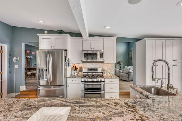 kitchen with light stone counters, stainless steel appliances, sink, white cabinets, and vaulted ceiling with beams