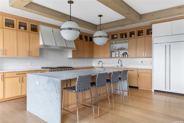 kitchen with light wood-type flooring, paneled fridge, wall chimney range hood, decorative light fixtures, and a center island with sink