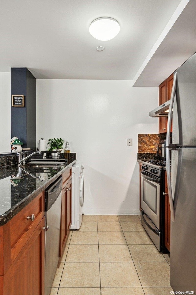 kitchen featuring exhaust hood, dark stone counters, appliances with stainless steel finishes, light tile patterned flooring, and washer / clothes dryer