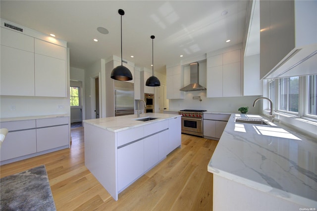 kitchen with appliances with stainless steel finishes, wall chimney exhaust hood, decorative light fixtures, a center island with sink, and white cabinets