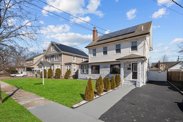 view of front facade featuring solar panels, fence, stone siding, a front lawn, and a chimney