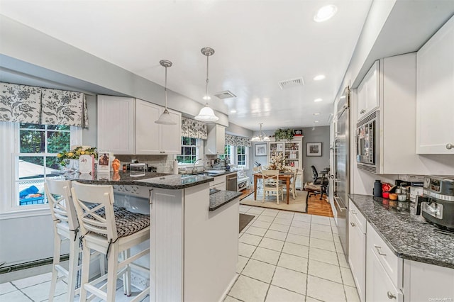 kitchen featuring a kitchen bar, dark stone counters, built in appliances, white cabinets, and hanging light fixtures