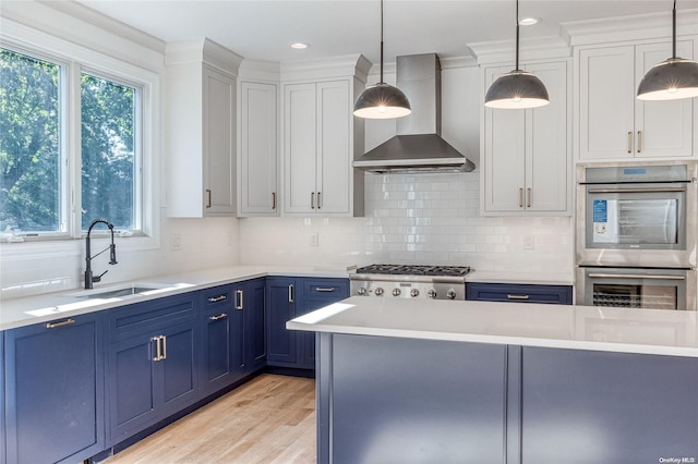 kitchen featuring wall chimney range hood, blue cabinetry, appliances with stainless steel finishes, decorative light fixtures, and white cabinetry