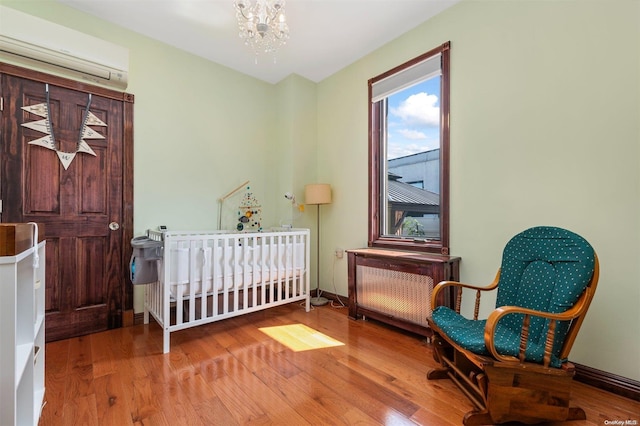 bedroom featuring a crib, hardwood / wood-style flooring, an AC wall unit, a chandelier, and radiator heating unit