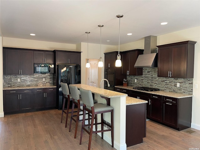 kitchen with a kitchen island with sink, black appliances, wall chimney range hood, wood-type flooring, and pendant lighting