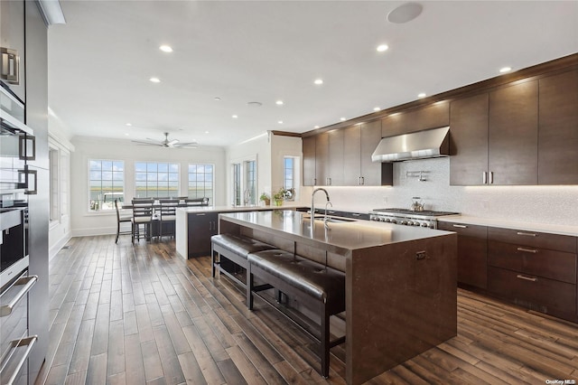 kitchen with sink, a kitchen island with sink, dark wood-type flooring, and wall chimney range hood