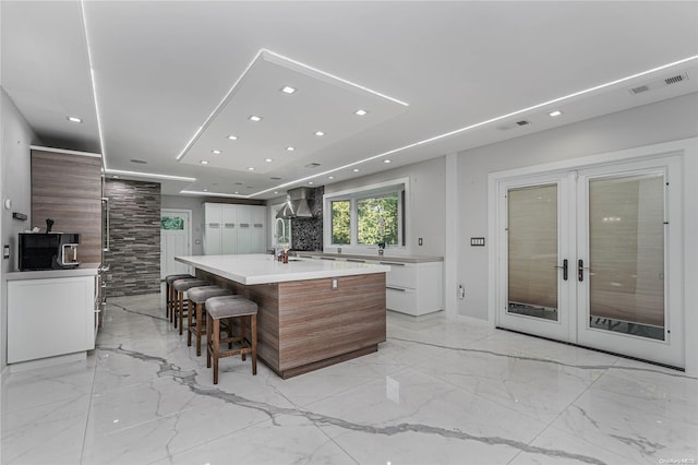 kitchen featuring a large island, french doors, wall chimney range hood, a kitchen bar, and white cabinets