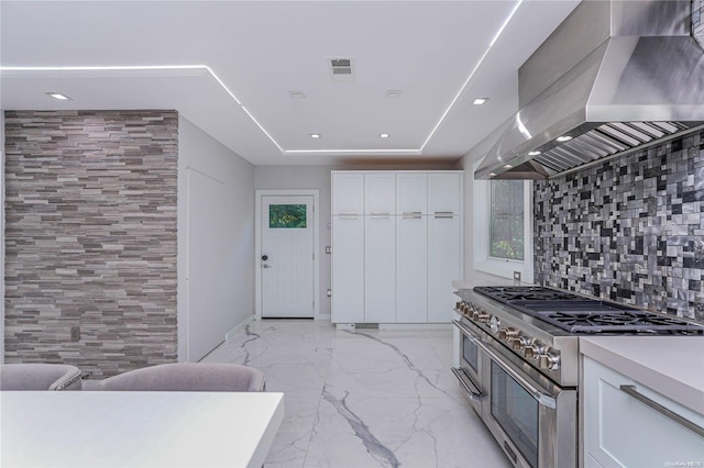 kitchen featuring wall chimney range hood, tasteful backsplash, white cabinets, and range with two ovens