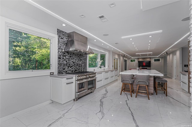 kitchen featuring wall chimney exhaust hood, a kitchen island with sink, range with two ovens, white cabinetry, and plenty of natural light