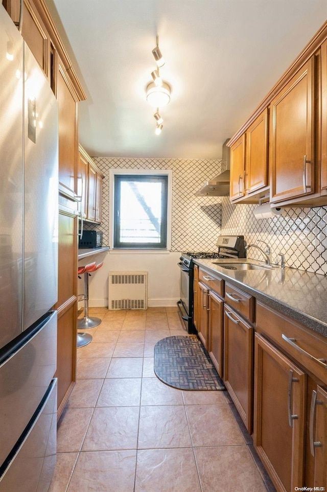 kitchen with wall chimney exhaust hood, gas range oven, tasteful backsplash, radiator heating unit, and stainless steel refrigerator