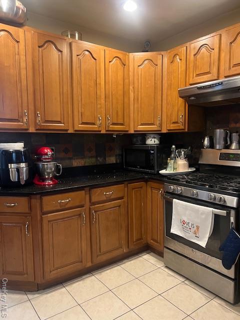 kitchen featuring gas range, under cabinet range hood, dark stone counters, and brown cabinetry