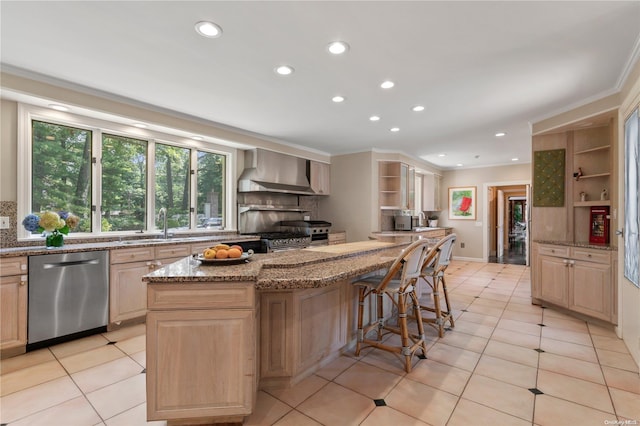 kitchen featuring light stone countertops, light brown cabinets, a center island, wall chimney range hood, and appliances with stainless steel finishes