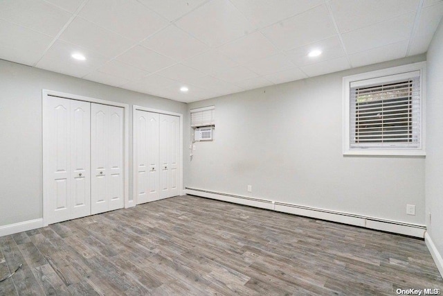 unfurnished bedroom featuring a paneled ceiling, a wall mounted air conditioner, two closets, baseboard heating, and wood-type flooring