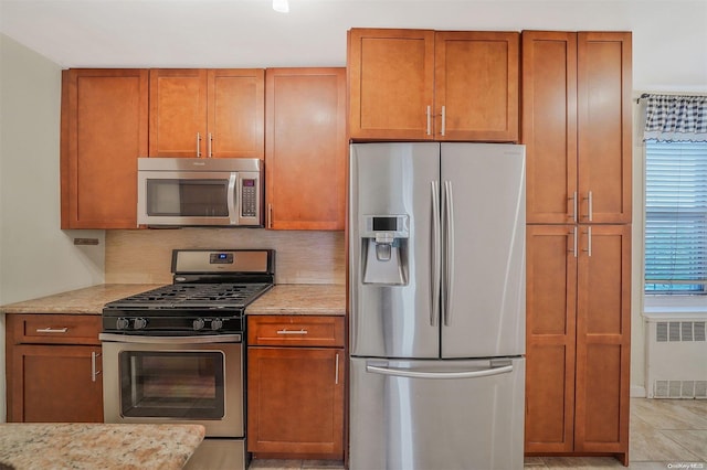 kitchen with decorative backsplash, appliances with stainless steel finishes, light tile patterned flooring, light stone counters, and radiator heating unit