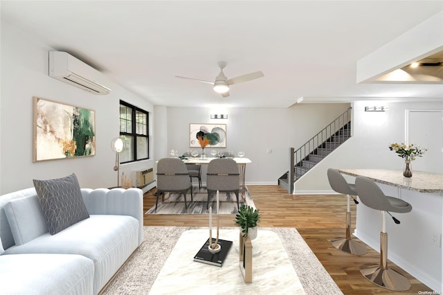 living room featuring a wall mounted air conditioner, light wood-type flooring, a ceiling fan, stairway, and baseboards