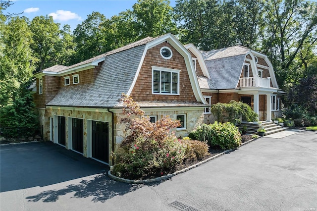 view of front of property featuring stone siding, aphalt driveway, a balcony, and a gambrel roof