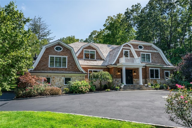 shingle-style home featuring a balcony, stone siding, driveway, and a gambrel roof