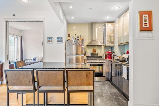 kitchen featuring decorative backsplash, appliances with stainless steel finishes, dark stone counters, wall chimney range hood, and a breakfast bar area