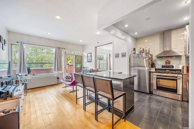 kitchen featuring hardwood / wood-style floors, wall chimney range hood, dark stone countertops, appliances with stainless steel finishes, and a kitchen bar