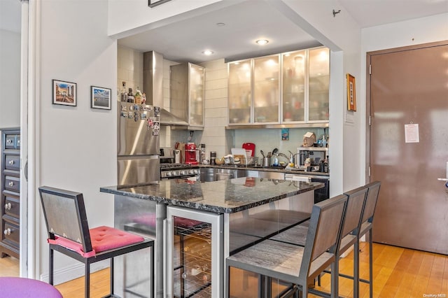 bar with stainless steel refrigerator, wall chimney exhaust hood, beverage cooler, dark stone countertops, and light hardwood / wood-style floors