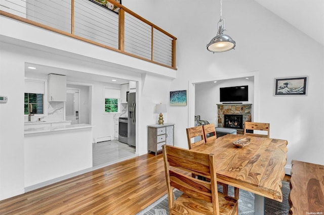 dining area featuring a baseboard heating unit, light wood-type flooring, high vaulted ceiling, and a stone fireplace