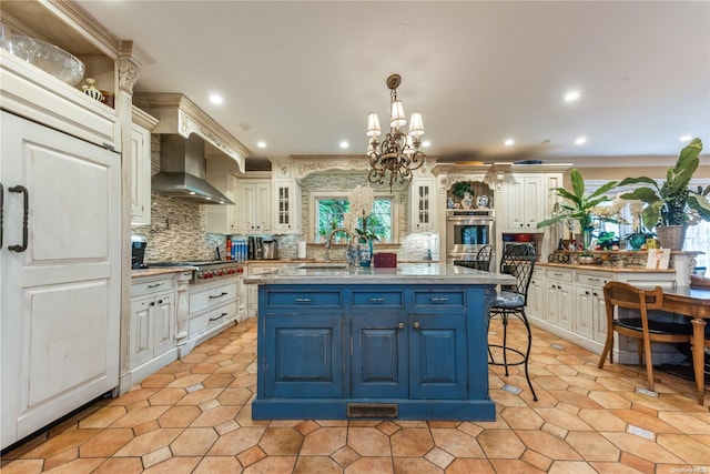 kitchen featuring wall chimney range hood, an inviting chandelier, decorative light fixtures, a center island with sink, and ornamental molding