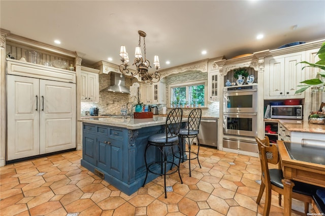 kitchen with tasteful backsplash, wall chimney exhaust hood, stainless steel appliances, decorative light fixtures, and an island with sink