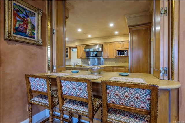 kitchen featuring stainless steel microwave, wall chimney exhaust hood, kitchen peninsula, crown molding, and decorative backsplash