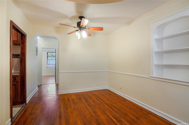 empty room with ceiling fan, built in features, ornamental molding, and dark wood-type flooring